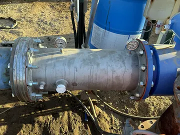 Large flanged stainless steel injector installed inline on an irrigation main beside a water tank