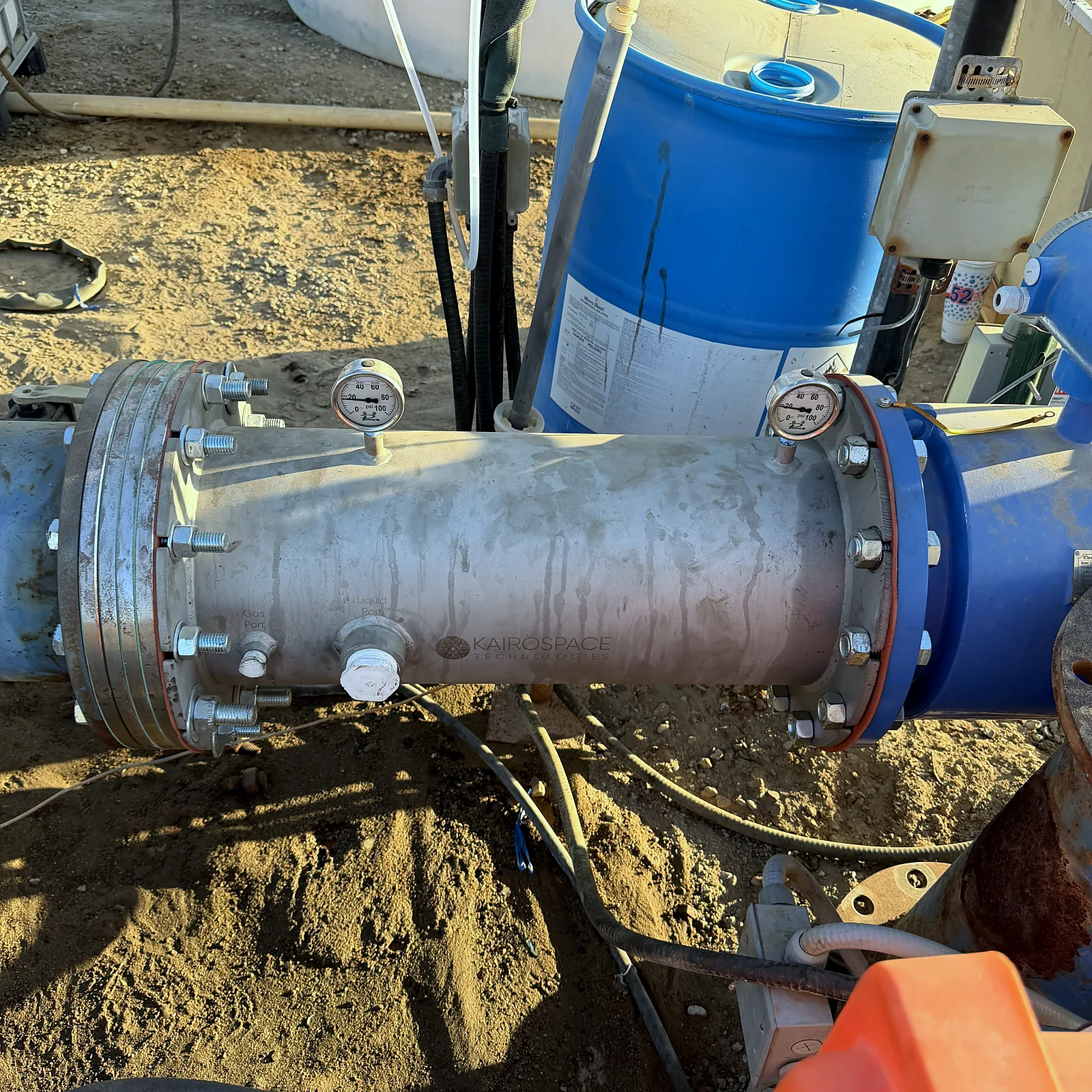 Large flanged stainless steel injector installed inline on an irrigation main beside a water tank, pressure gauges at both ends