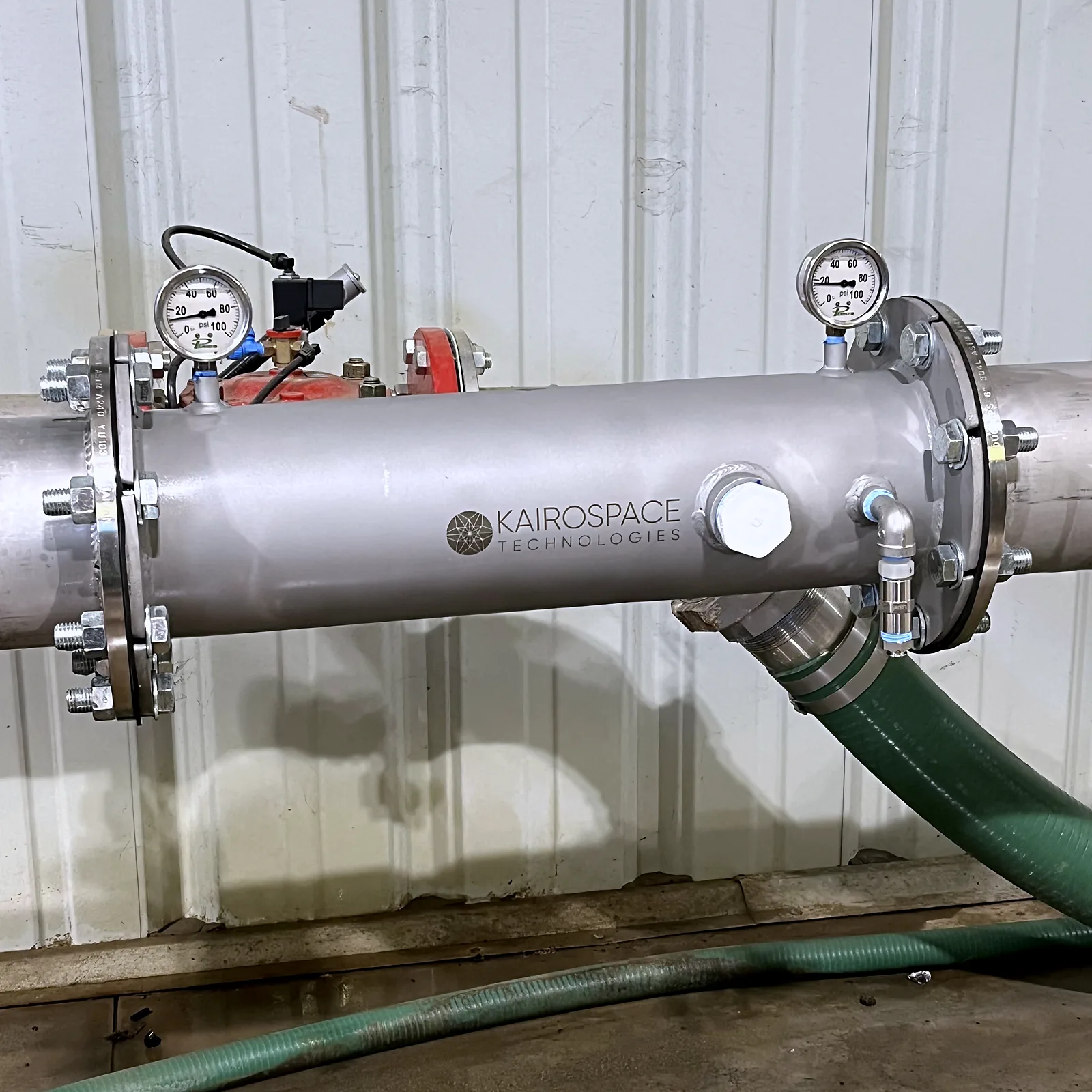 Stainless injector mounted horizontally inside a pump house with a gauge at each flange and a green suction hose running from the discharge