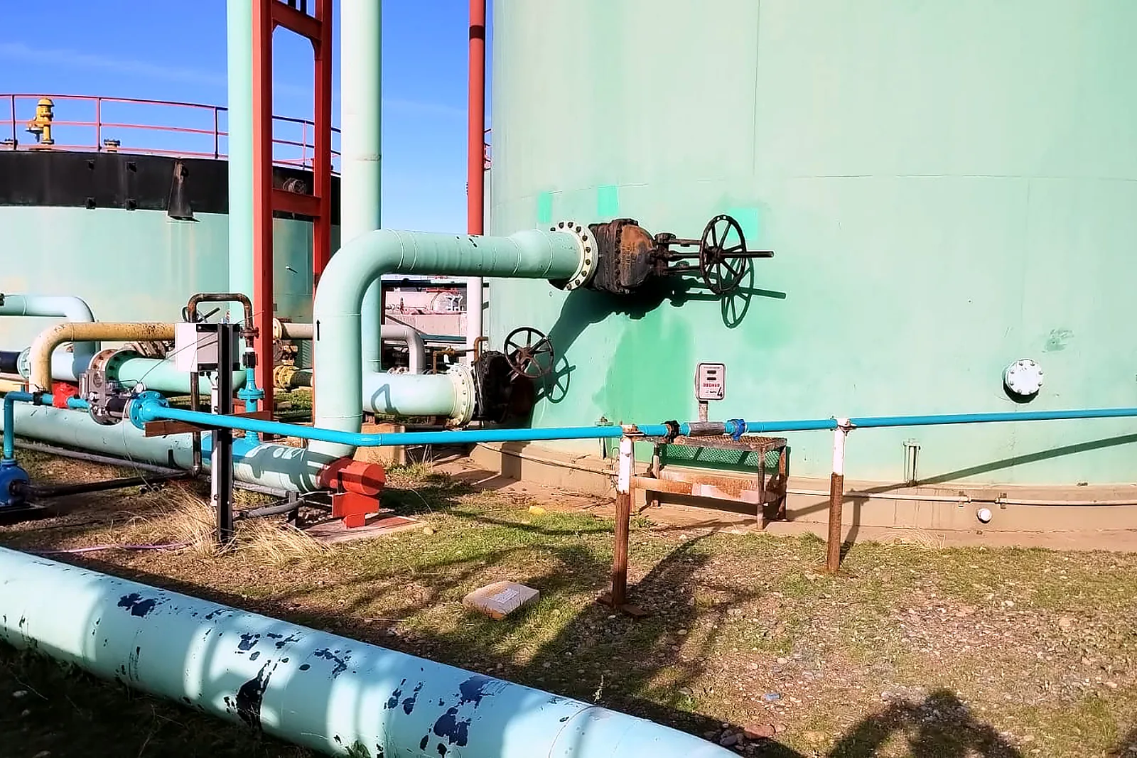 Blue injection line running along the base of a large storage tank and tying into the process piping at a tank battery